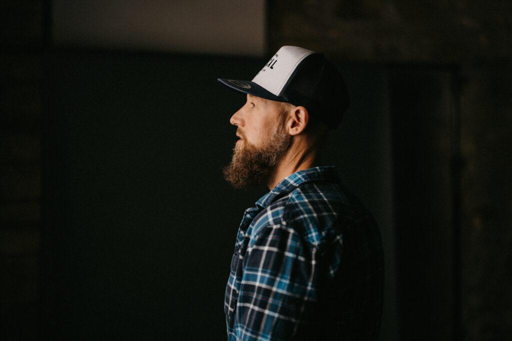 Side profile portrait of a bearded man in a cap and plaid shirt Bearded man wearing a baseball cap and plaid shirt, shown in side profile against a dark, moody background.
